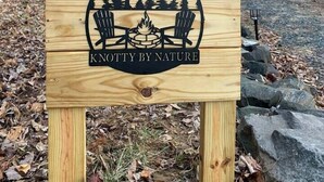 A wooden sign with a campfire, chairs, and trees, labeled "KNOTTY BY NATURE."