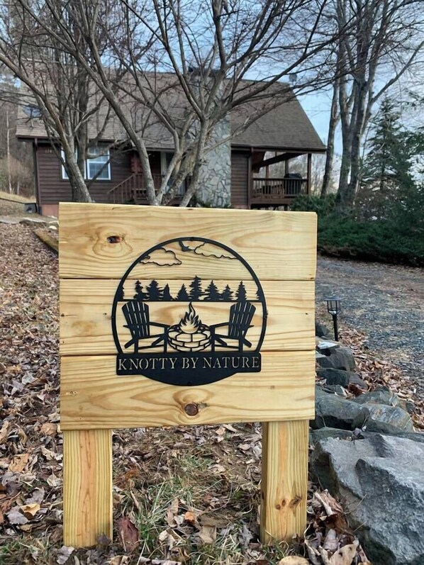 A wooden sign with a campfire, chairs, and trees, labeled "KNOTTY BY NATURE."