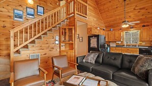 A wooden cabin interior with a staircase, a black leather sofa, two leather armchairs, a coffee table, and a kitchen area.