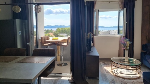 Un salon moderne avec un coin repas, de grandes fenêtres avec des rideaux, une table basse ronde en verre et une vue sur l’extérieur.