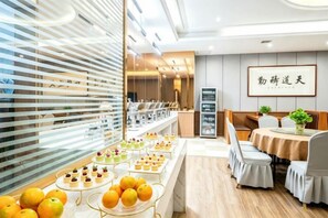 Un restaurant doté d’un long comptoir où sont exposés des desserts et des fruits, d’un espace avec des tables et des chaises, et d’un mur orné d’une enseigne encadrée.