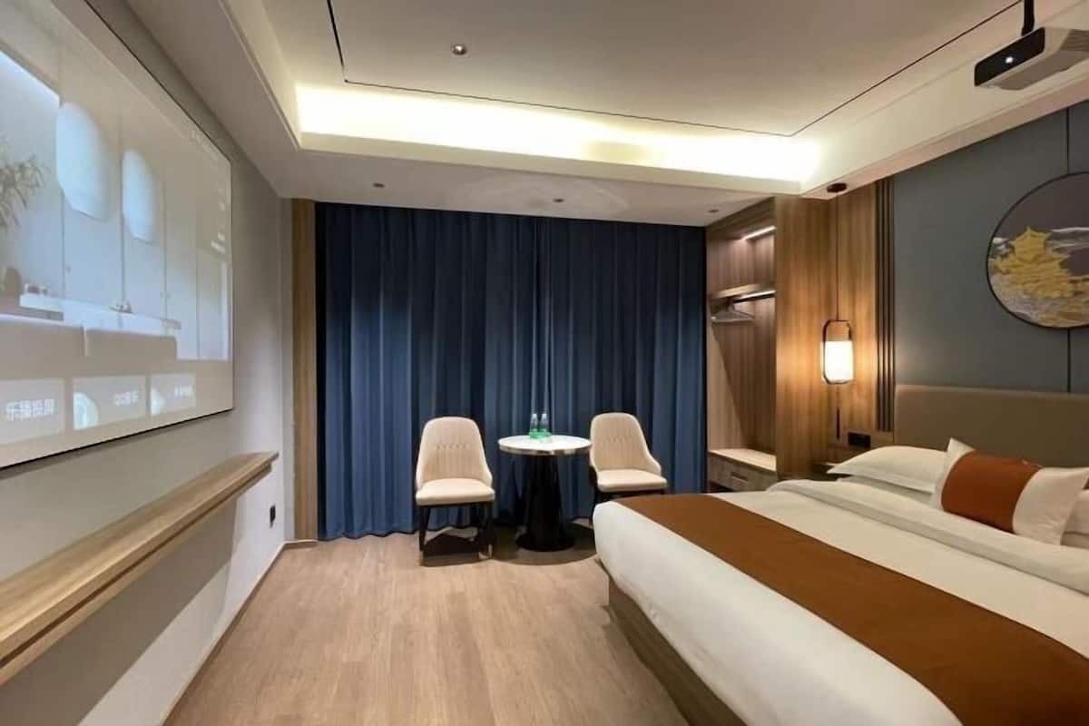 Et hotelværelse med seng, to stole, et lille bord, et fjernsyn og en vægmonteret projektor.