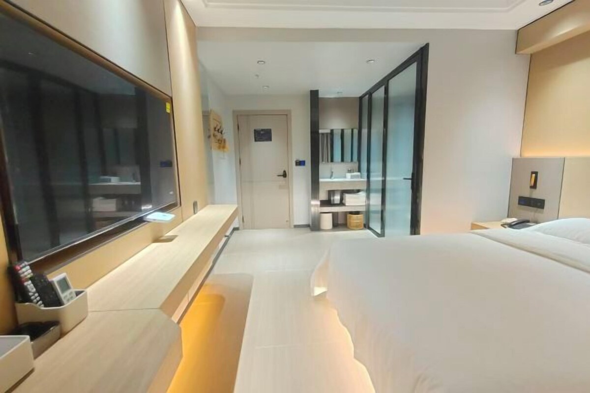A modern hotel room with a large bed, a flat-screen TV, a bedside table with a lamp, and a bathroom with a glass shower enclosure.