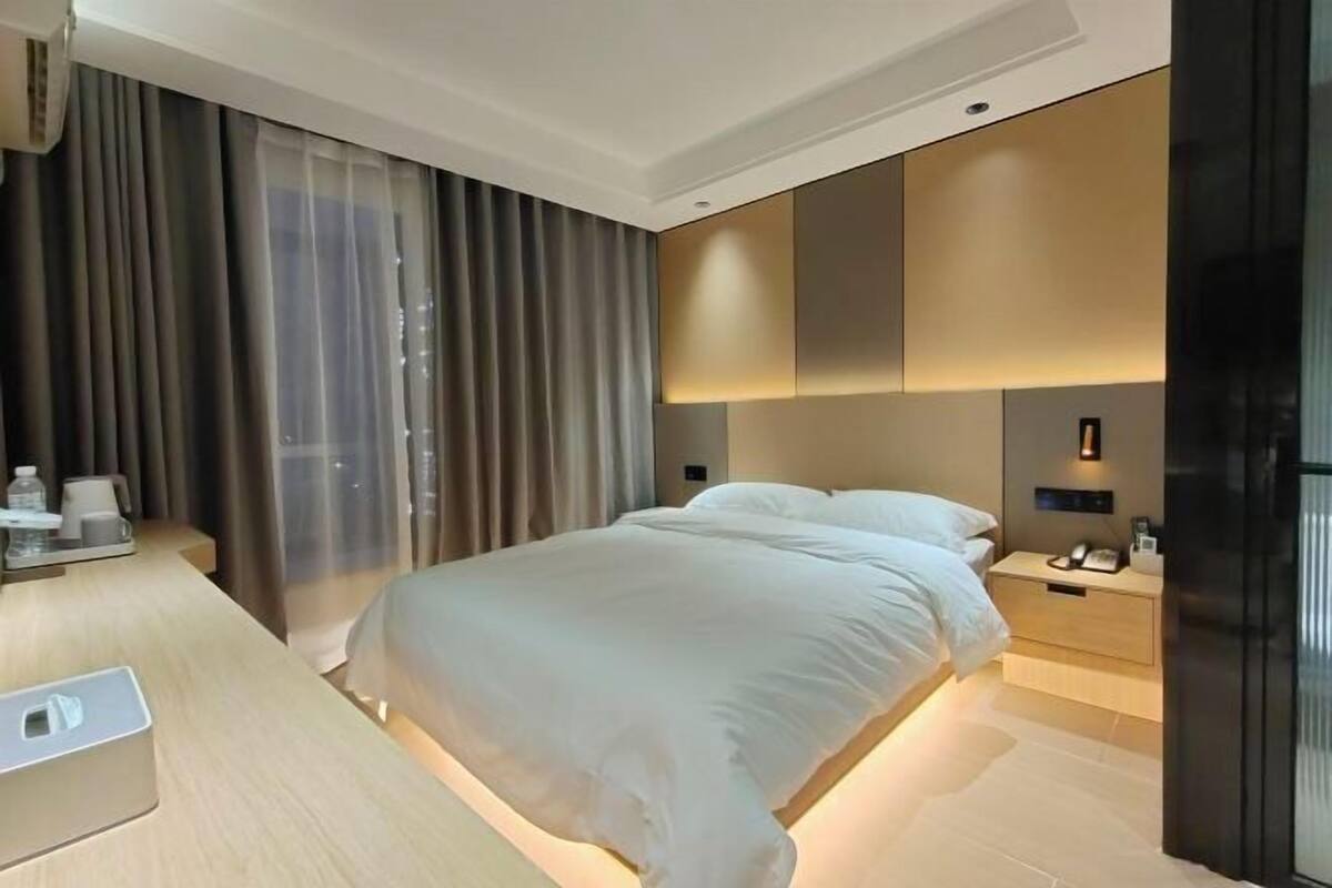 Une chambre d’hôtel moderne avec un lit bien fait, une table de chevet avec une lampe et une grande fenêtre avec des rideaux.