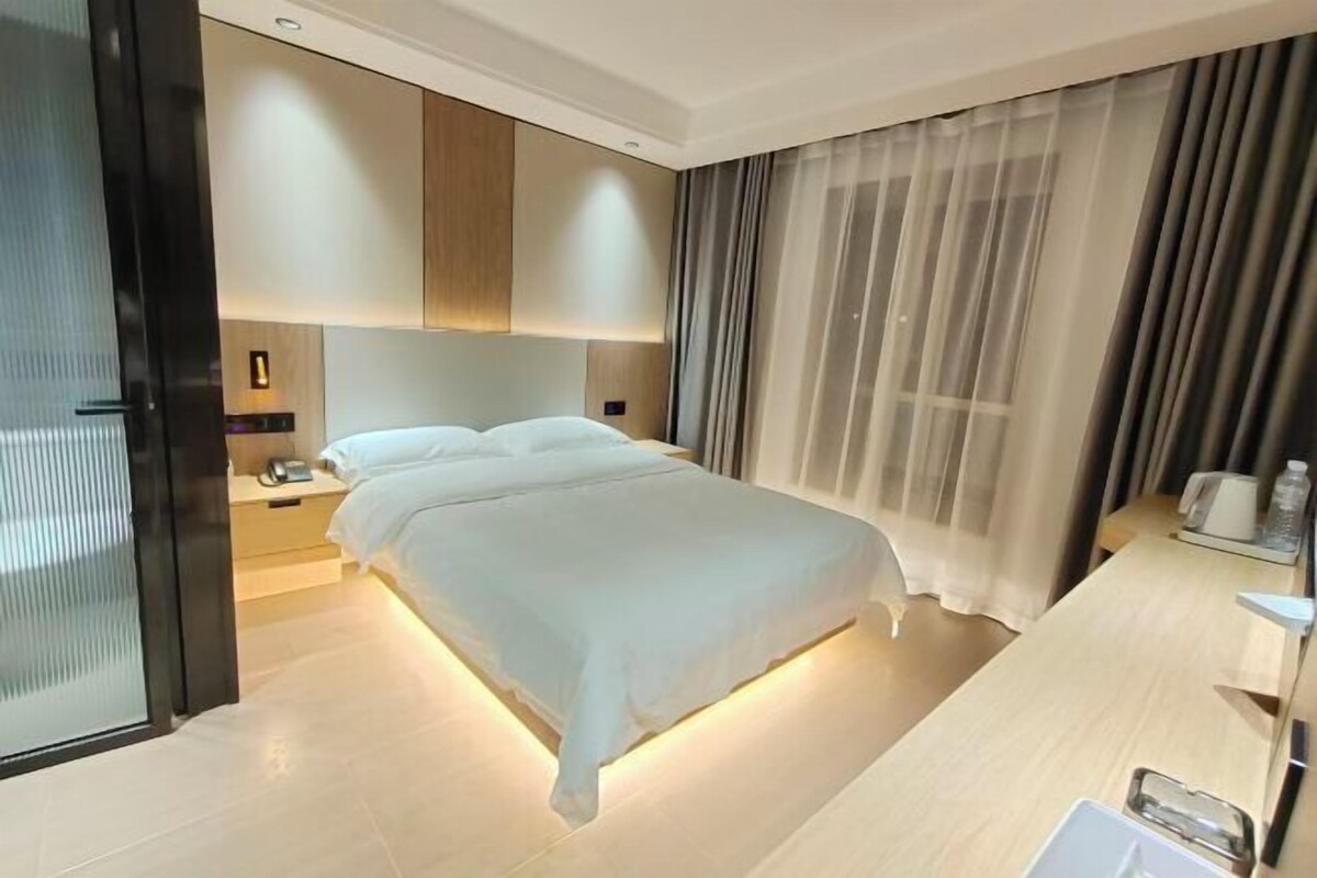 A modern hotel room with a large bed, a bedside table with a lamp, and a view of the city through sheer curtains.