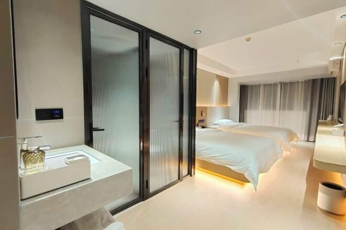 Une chambre d’hôtel moderne dotée d’une grande porte-fenêtre coulissante, d’une salle de bain séparée avec un lavabo, de deux lits et d’une table de chevet avec une lampe.