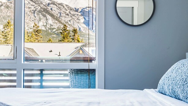 A bedroom with a large window, a round mirror, and a bed with white bedding. - "Tranquil Tamarack" at Tamarack Lodge (Canmore)