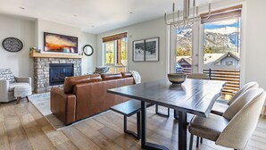 A modern living room with a fireplace, a dining area with a large table, and a view of mountains through large windows. - "Tranquil Tamarack" at Tamarack Lodge (Canmore)