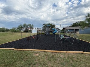 A playground with a slide and swings, surrounded by a grassy area and a building in the background.