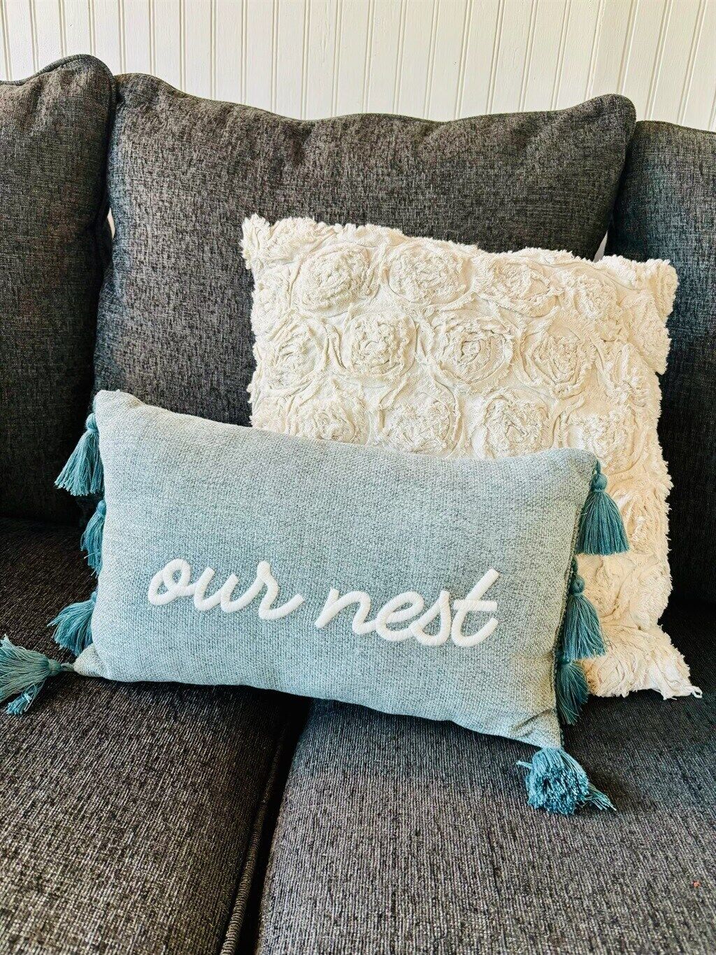 Un canapé de couleur sombre agrémenté de trois coussins décoratifs. L’un des coussins présente un motif floral, un autre est orné de glands, et le troisième est brodé de texte.