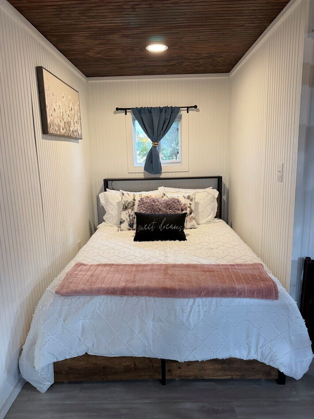 Un dormitorio con cama, ventana con cortinas, luz de techo e imagen colgada en la pared.