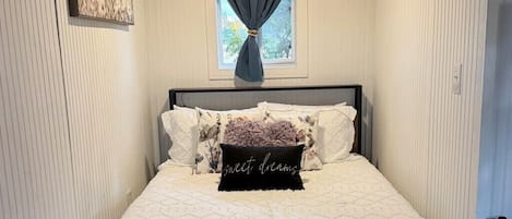 Un dormitorio con cama, ventana con cortinas, luz de techo e imagen colgada en la pared.