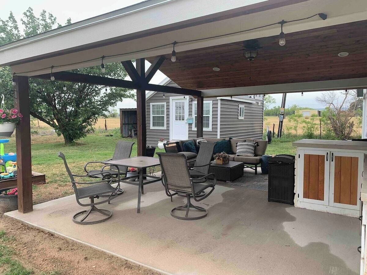 A covered patio with outdoor furniture, a grill, and a small shed in the background.