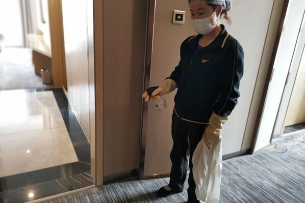 A person in a uniform standing in a hallway with a door, holding a bag and a phone. - New Times Inn & Hotel (Tongchuan)
