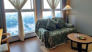 A room with a floral-patterned sofa, a small round table with a lamp, and a view of a landscape through large windows with sheer curtains. - New Times Inn & Hotel (Tongchuan)