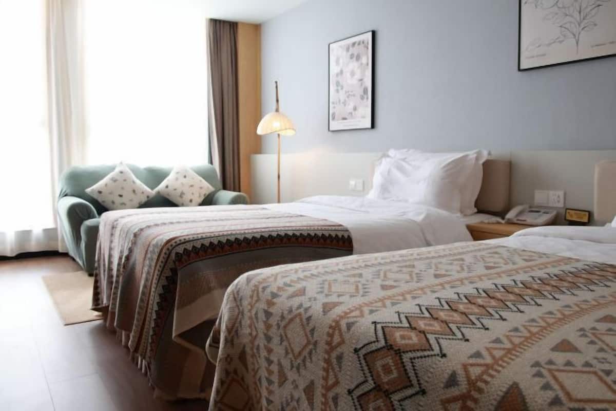 Une chambre d’hôtel avec deux lits, un fauteuil vert, une lampe et une estampe botanique encadrée au mur.
