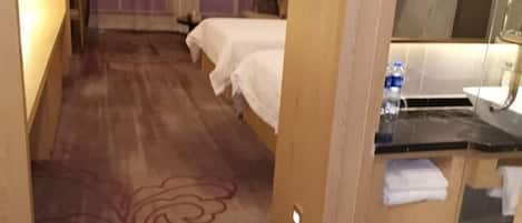 A hotel room with two beds, a bathroom with a mirror and sink, and a wooden floor.