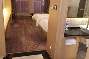 A hotel room with two beds, a bathroom with a mirror and sink, and a wooden floor. - New Times Inn & Hotel (Tongchuan)