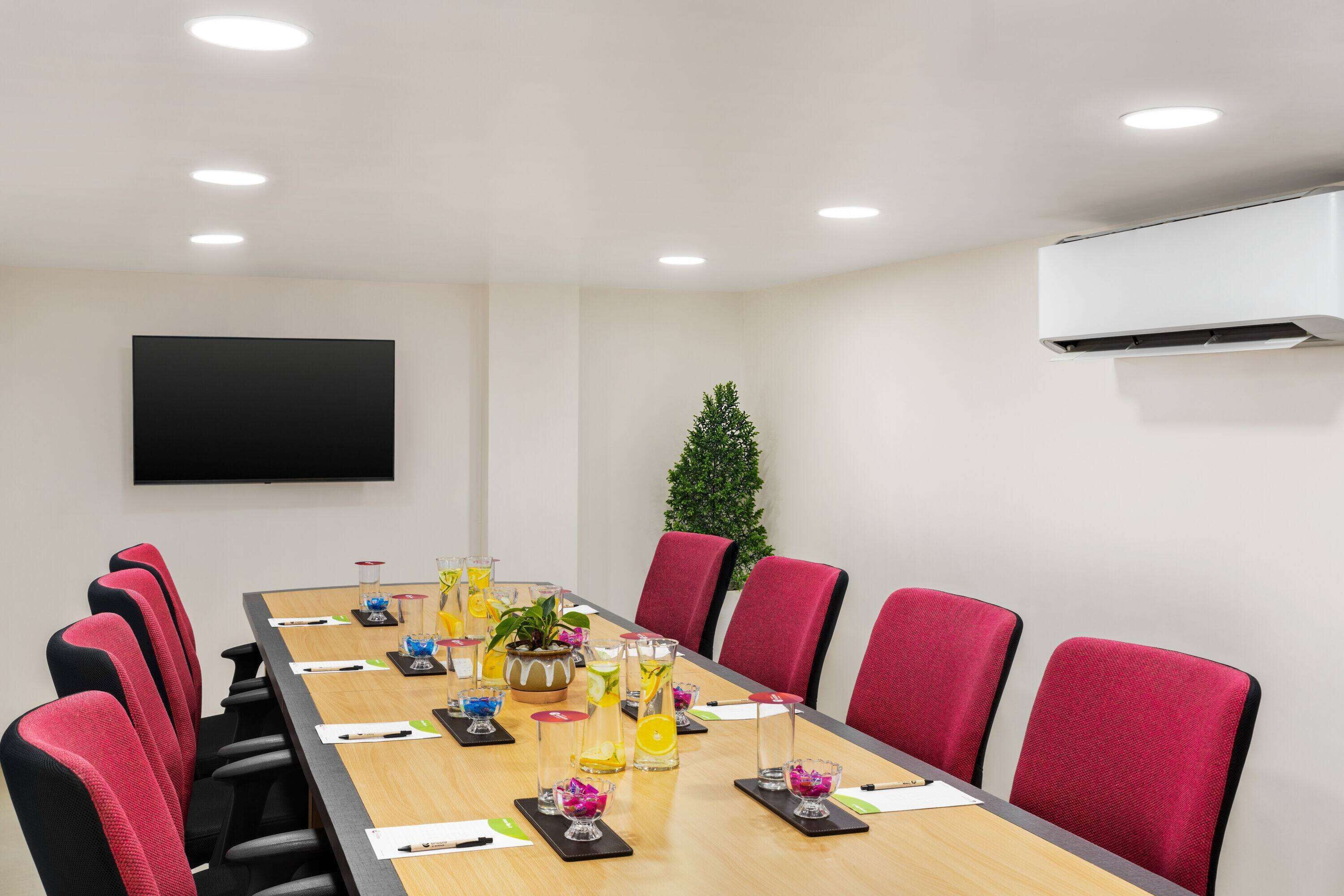 A conference room with a long table, red chairs, a flat-screen TV, and an air conditioning unit.