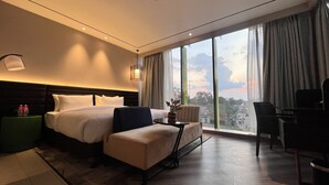 A hotel room with a large bed, a sofa, a desk, and a view of the city through large windows. - Ramada Encore By Wyndham Bareilly Civil Lines (Bareilly)