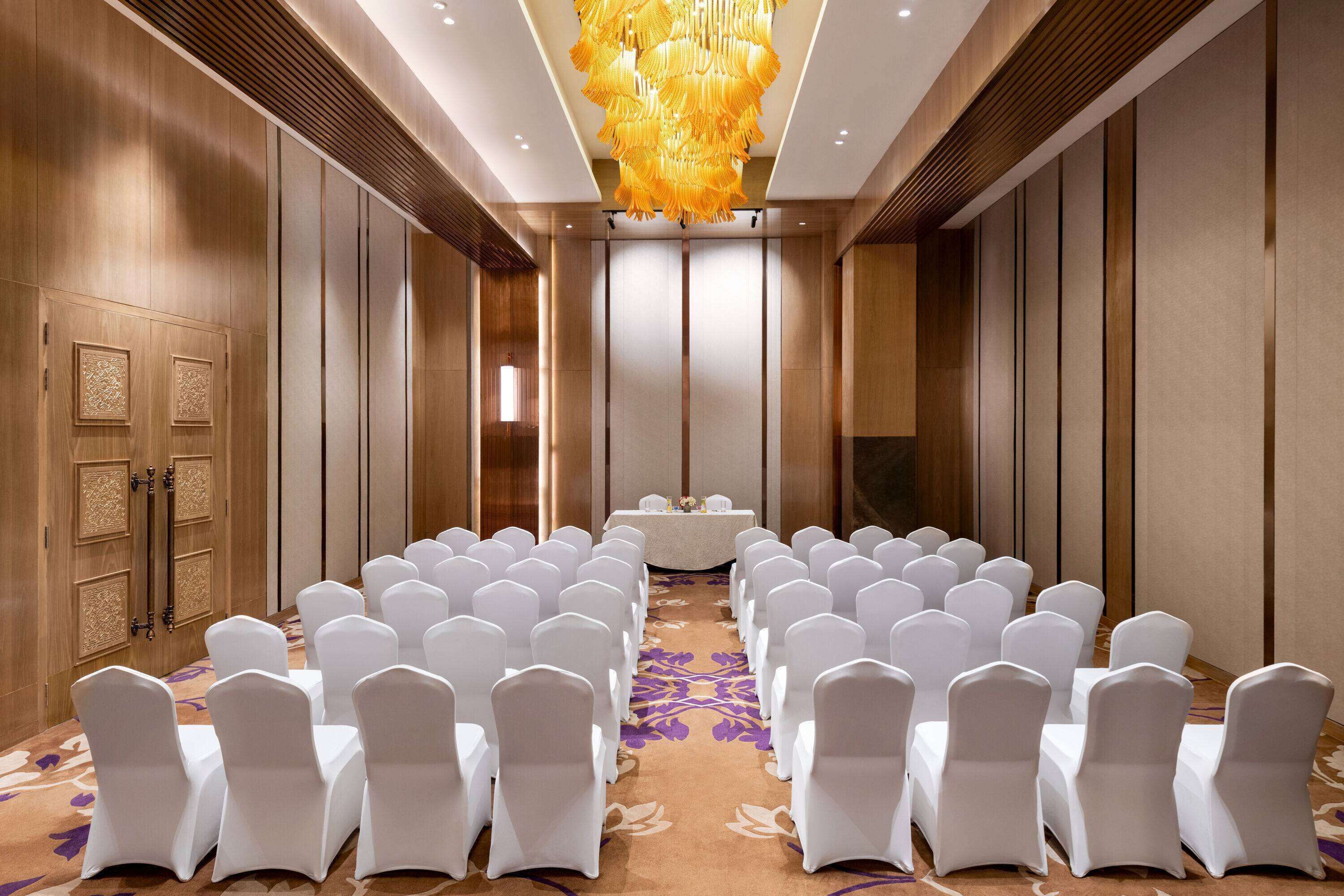 A conference room with rows of white chairs, a central table, and a decorative chandelier.