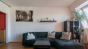 A modern living room with a dark green sectional sofa, a black coffee table, a painting of trees on the wall, a small shelf with candles and flowers, and a bookshelf with plants. - Cosy, sunny two-story apartment, 20min center (Hlavní mesto Praha)