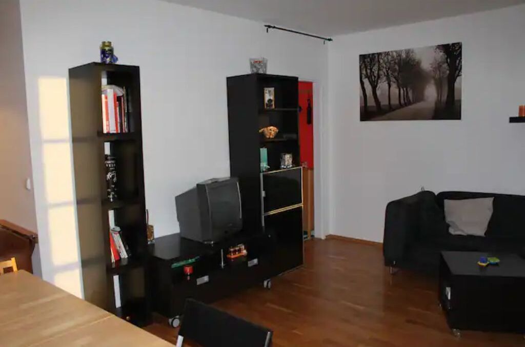 A living room with a wooden floor, a black sofa, a coffee table, and two black shelving units with various items.