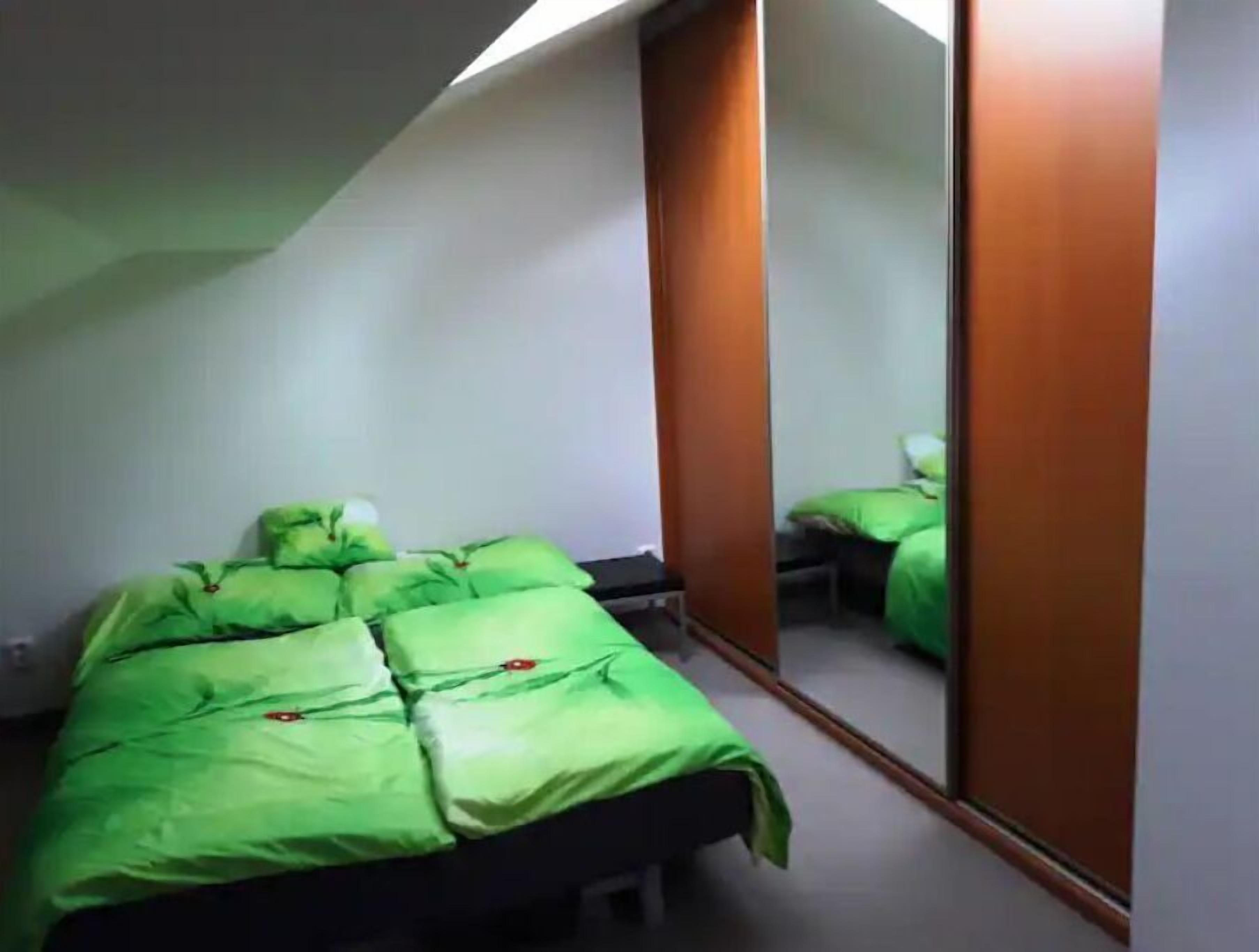 Une petite chambre avec un lit simple, dotée de draps verts et d’un grand miroir sur un des murs.