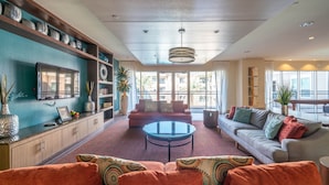 A spacious living room with a sofa, armchairs, a coffee table, and a TV on the wall. There are shelves with decorative items and a large window providing natural light.