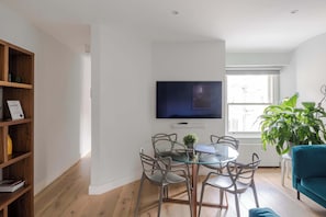 A modern living room with a dining table, chairs, a television, a bookshelf, and a large window.