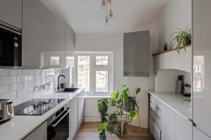 Une cuisine moderne avec des meubles blancs, un micro-ondes noir, un évier et une fenêtre donnant sur les bâtiments environnants.