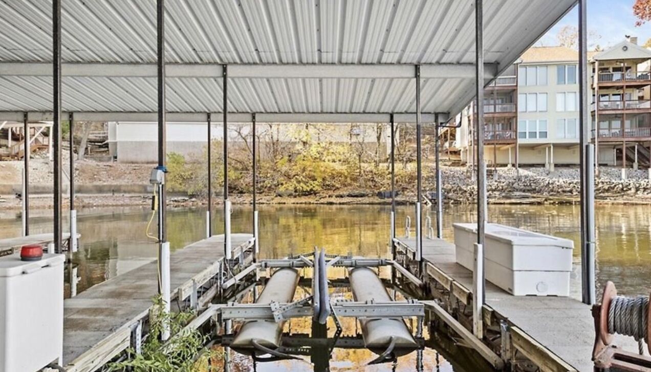 Un muelle techado con una masa de agua al fondo, estructuras metálicas y un edificio con balcones.