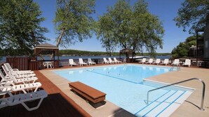 Ein Schwimmbecken mit Liegestühlen und einer Holzterrasse, umgeben von Bäumen und Blick auf einen See.