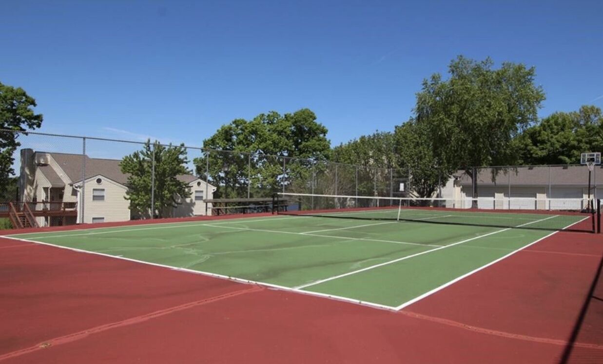 A tennis court with a net, surrounded by a fence, and adjacent buildings.