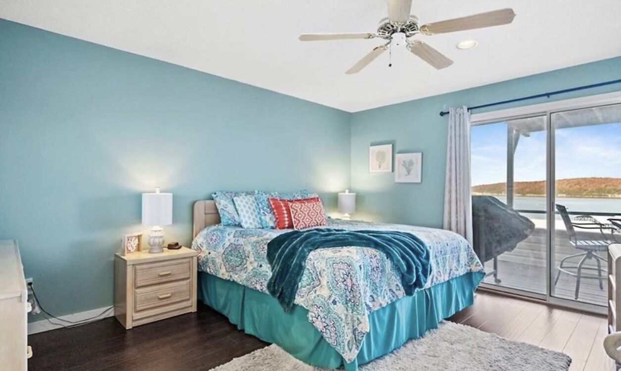 A bedroom with a large bed, a ceiling fan, a nightstand with a lamp, and a view of the outdoors through a sliding glass door.