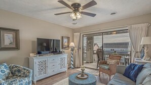 A living room with a ceiling fan, a television, a white cabinet, a blue patterned sofa, a round coffee table, and a view through a glass door.