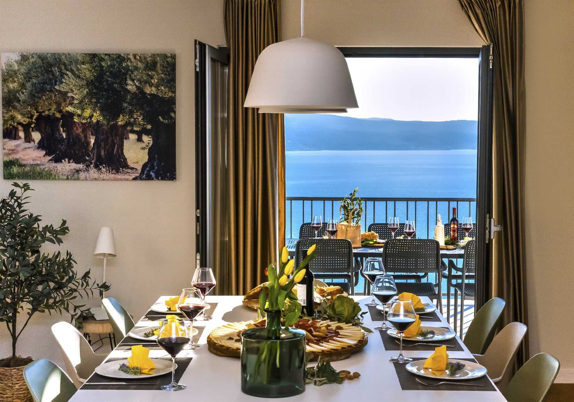 Mesa de comedor lista para cuatro personas con vista al mar, un cuadro de árboles y una lámpara colgante.