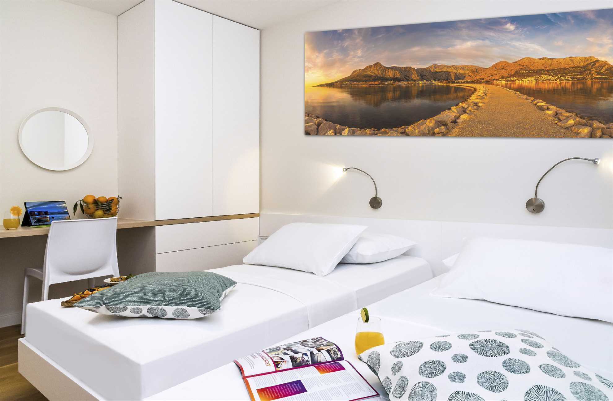 A hotel room with a bed, a desk, a chair, a round mirror, and a scenic landscape painting on the wall.