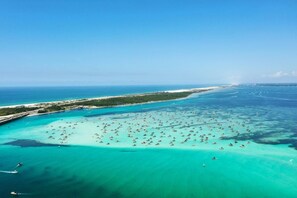 Vista aérea de una zona costera con numerosos barcos anclados en aguas turquesas cristalinas, un muelle y una costa distante con edificios.