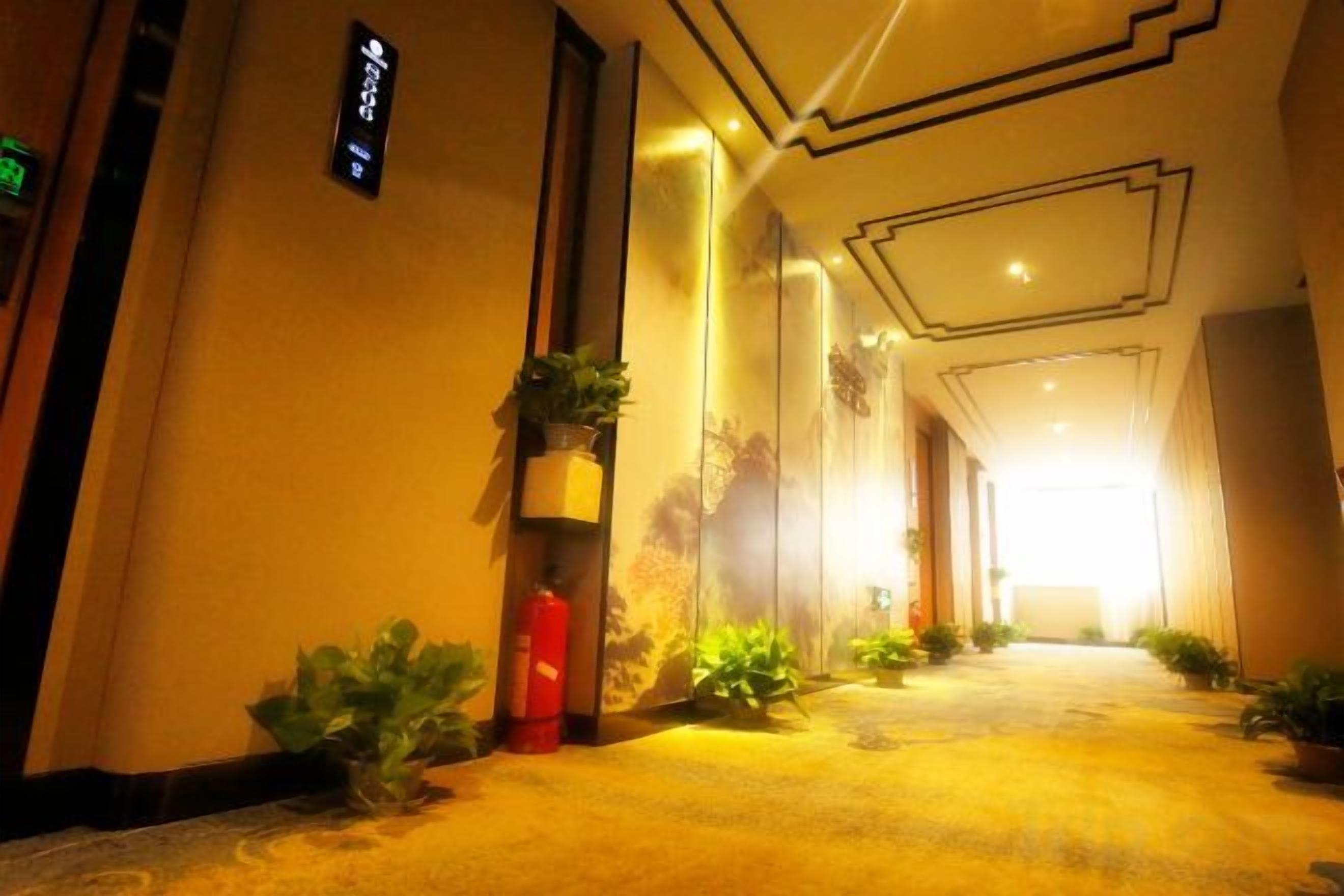 Un pasillo de hotel con piso alfombrado, plantas en macetas y un extintor.
