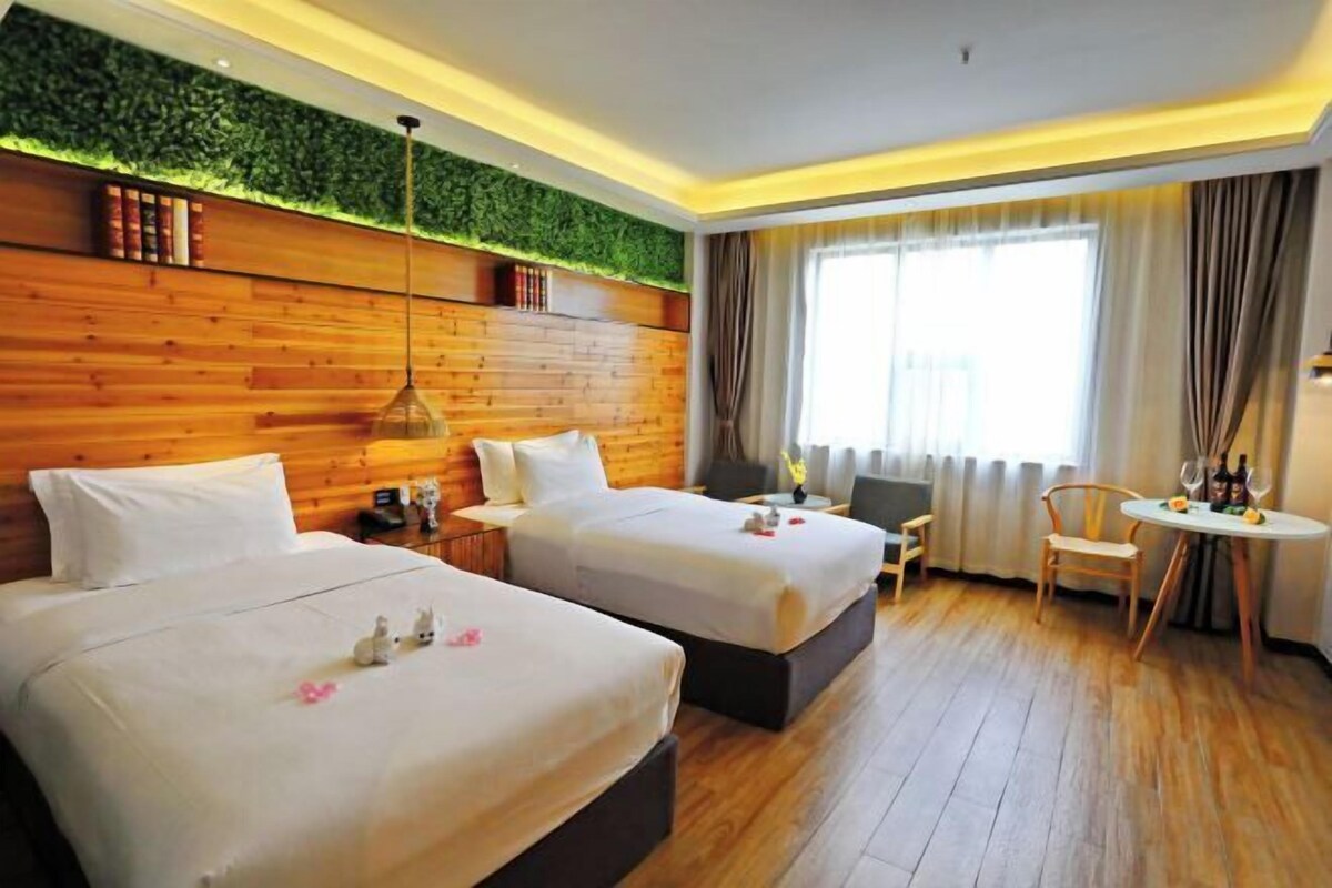 A hotel room with two beds, a wooden headboard, a green wall, a large window with curtains, a small table with wine glasses, and a chair.
