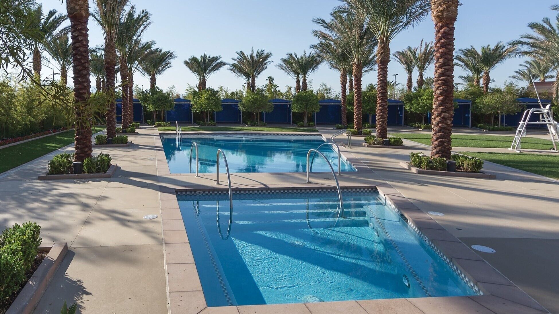 A rectangular swimming pool with two diving boards, surrounded by palm trees and a covered area in the background.