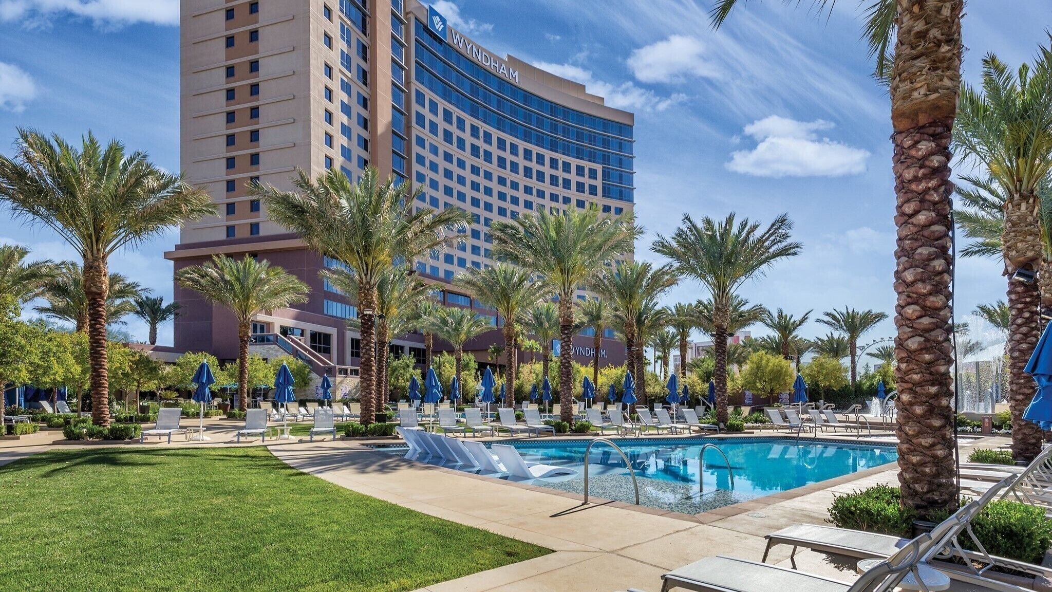 A hotel pool area with lounge chairs and umbrellas, palm trees, and a large hotel building in the background.