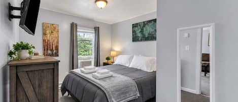 A bedroom with a bed, a TV mounted on the wall, a window with curtains, and a plant on a side table.