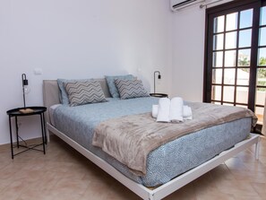 A single-bed room with a patterned bedspread, two pillows, a bedside table with a lamp, and a window with a view.