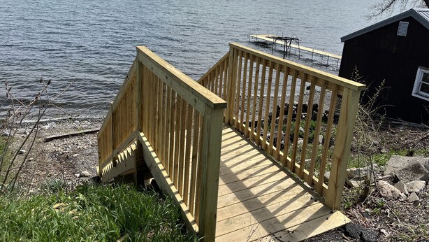 Eine Holzterrasse führt zu einem kleinen Steg am See, mit einem Gebäude im Hintergrund.