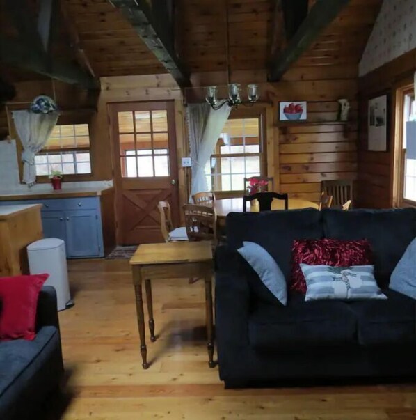 A cozy cabin interior with a wooden floor, a black sofa with cushions, a dining table, and a kitchen area with wooden cabinets and a window. - Log Home Leisure (Romulus)