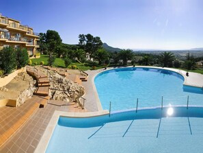 A large, curved swimming pool with a clear blue waterline, surrounded by a tiled deck and a stone pathway leading to a building with balconies. - APCOSTAS Club Torre Vella / One Bedroom (L'Estartit)