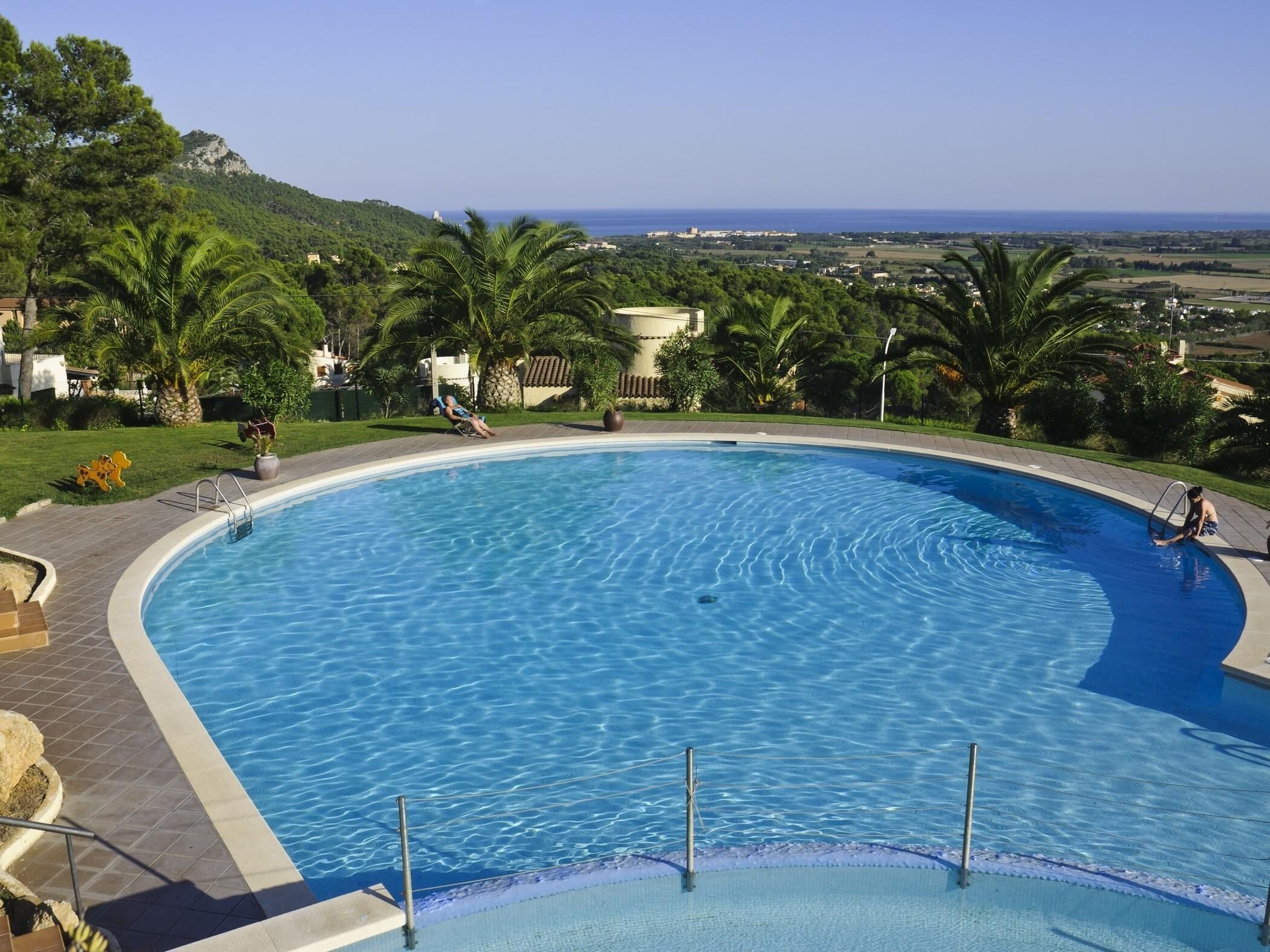 A large, curved swimming pool with clear blue water, surrounded by palm trees and a view of the sea and hills in the background.