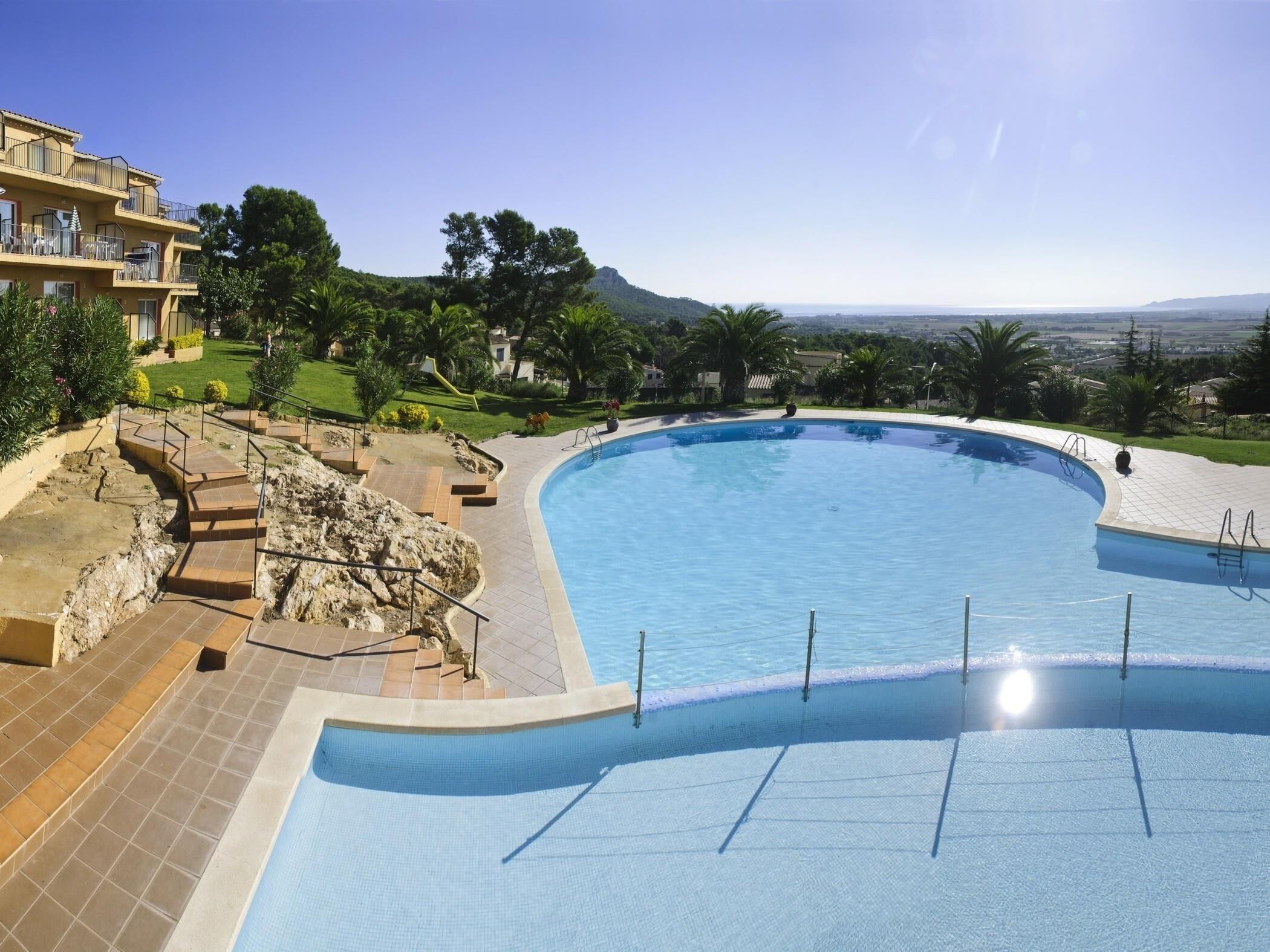 Ein großer, gewölbter Pool mit freiem Blick auf die umliegende Landschaft, darunter ein Gebäude mit Balkonen und ein ferner Gebirgszug.
